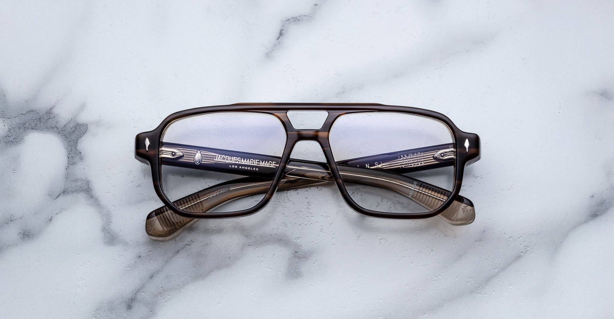 The Jacques Marie Mage Evans eyeglasses by Jacques Marie Mage feature a brown, oversized rectangular acetate frame, displayed on a white marble surface.