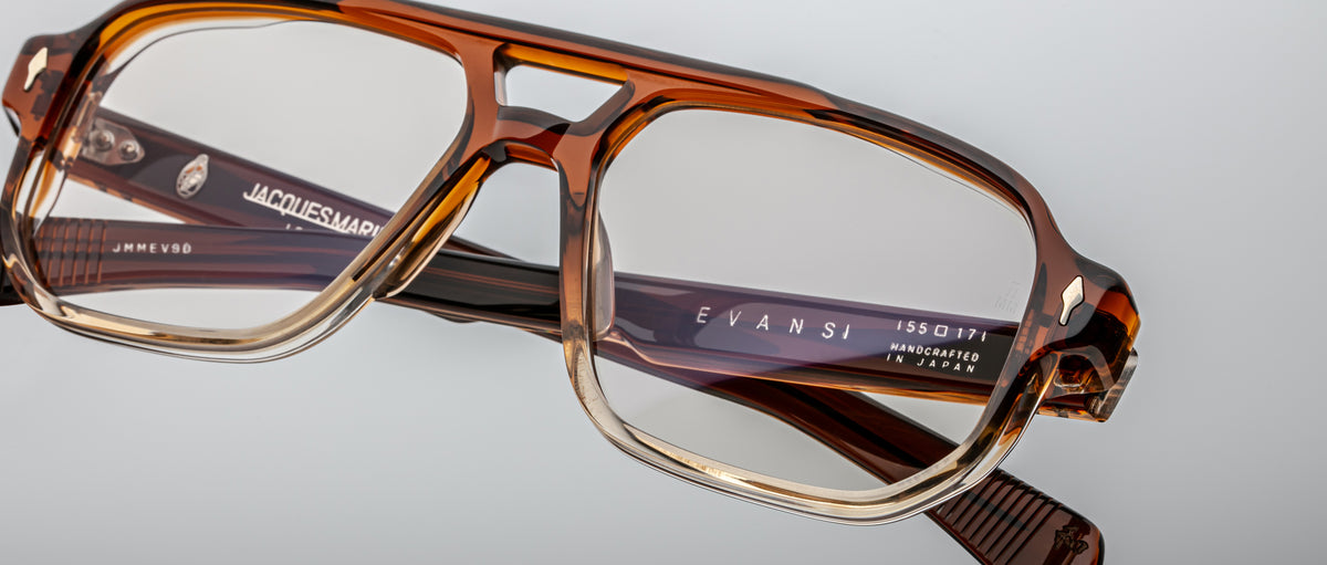 Close-up of handcrafted Jacques Marie Mage Evans optical glasses with a brown and clear oversized acetate frame, featuring the Jacques Marie Mage logo on the temples, set against a plain background.