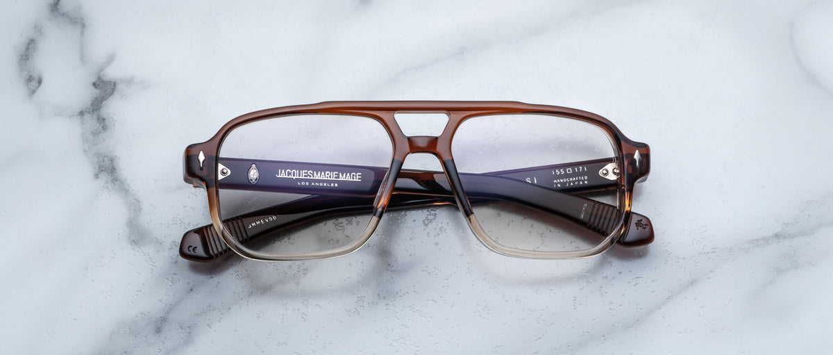 A pair of Jacques Marie Mage Evans glasses, featuring a brown oversized rectangular acetate frame with clear lenses, is displayed on a white marble surface.
