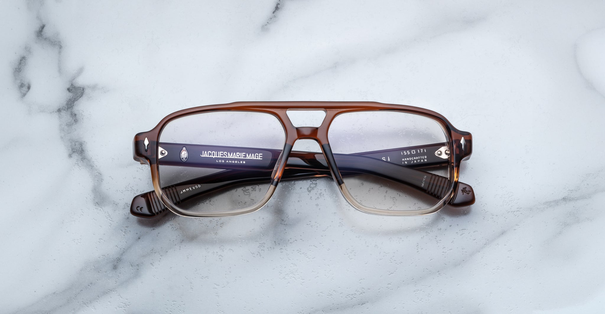 A pair of Jacques Marie Mage Evans glasses, featuring a brown oversized rectangular acetate frame with clear lenses, is displayed on a white marble surface.