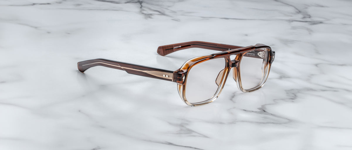 A pair of Jacques Marie Mage Evans eyeglasses with brown, square acetate frames and clear lenses rests on a white marble surface.