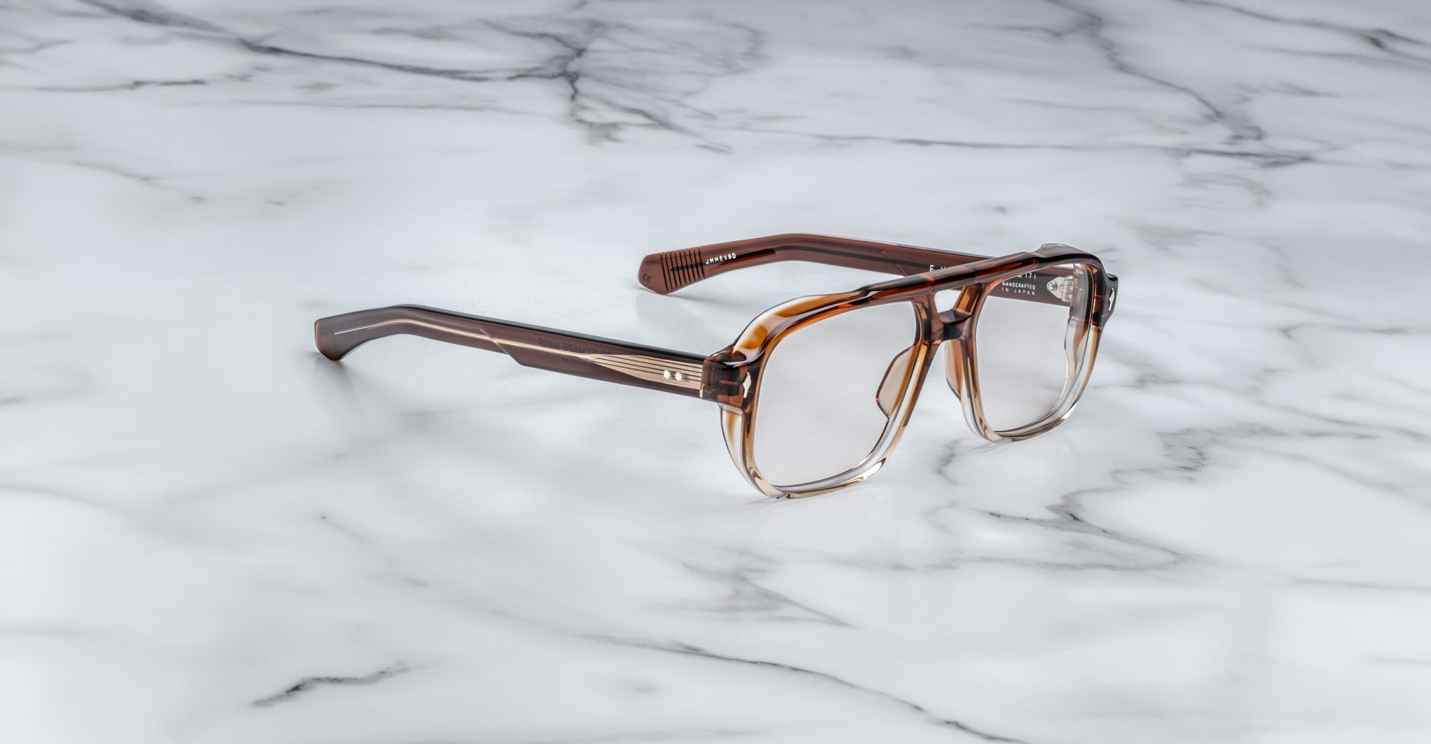 A pair of Jacques Marie Mage Evans eyeglasses with brown, square acetate frames and clear lenses rests on a white marble surface.