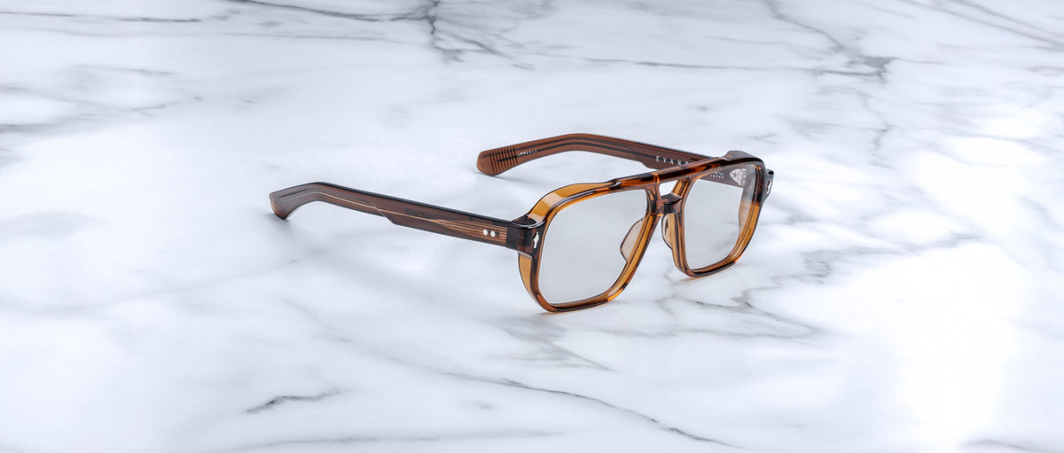 The Jacques Marie Mage Evans, a pair of oversized brown square acetate eyeglasses by Jacques Marie Mage, is displayed on a white marble surface.