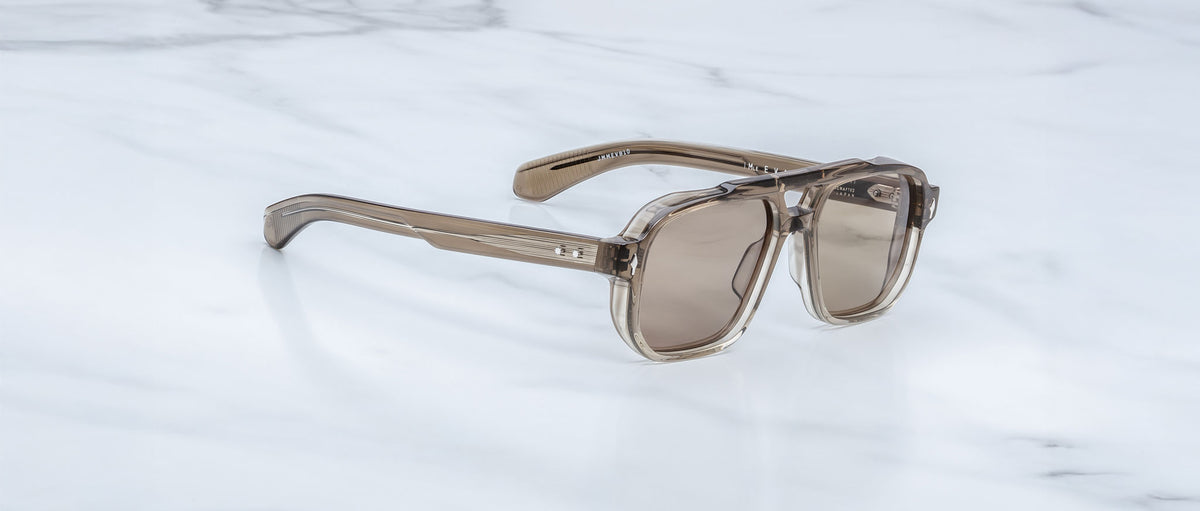 The Jacques Marie Mage Evans sunglasses feature translucent brown rectangular lenses and an oversized acetate frame, resting on a white marble surface and evoking the brand’s signature handcrafted optical style.