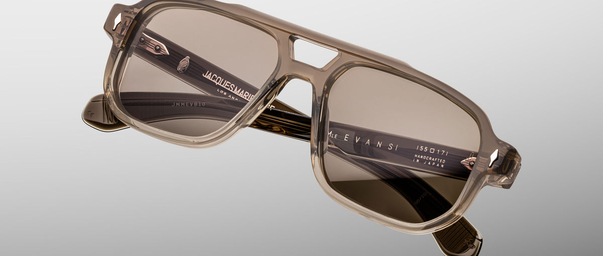 The Jacques Marie Mage Evans sunglasses feature a rectangular, taupe acetate oversized frame with dark tinted lenses, inspired by Jacques Marie Mage design, shown on a light gray background.