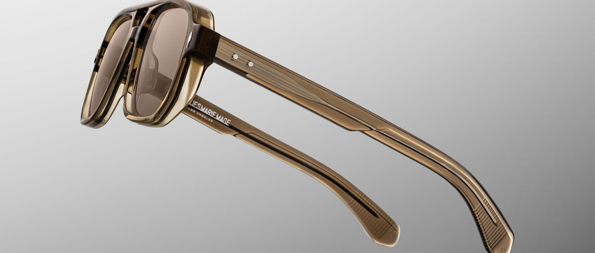 Close-up of Jacques Marie Mage Evans sunglasses, featuring an oversized brown translucent acetate frame and tinted lenses, displayed from a side angle against a gradient gray background, echoing the handcrafted quality of Jacques Marie Mage eyewear.