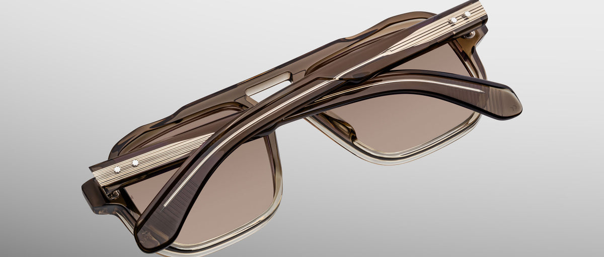 Jacques Marie Mage Evans sunglasses feature folded brown oversized acetate frames with gold temple accents, echoing the brand's signature handcrafted style, presented against a plain gradient background.