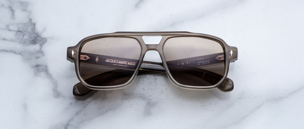The Jacques Marie Mage Evans sunglasses by Jacques Marie Mage feature an oversized brown acetate square frame with dark lenses, displayed on a white marble surface.