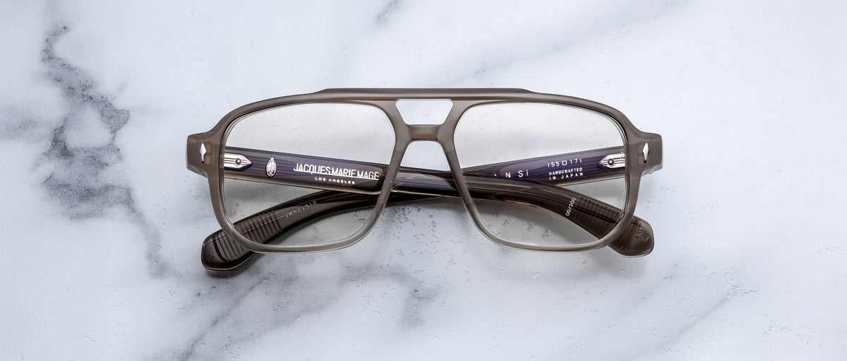 The Jacques Marie Mage Evans eyeglasses by Jacques Marie Mage feature an oversized gray acetate frame with thick rims and rectangular lenses, shown resting on a white marble surface.