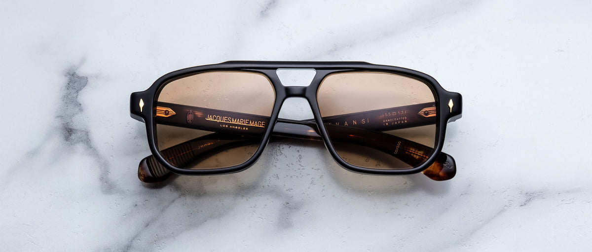 A pair of Jacques Marie Mage Evans sunglasses with brown-tinted, square oversized acetate frames rests on a white marble surface.