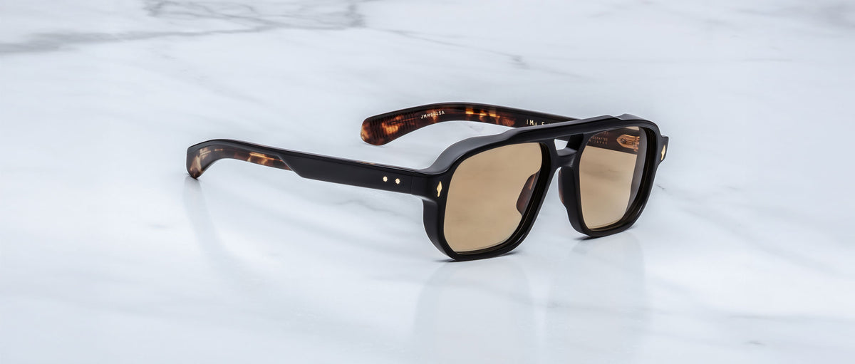 The Jacques Marie Mage Evans sunglasses in Noir 15 feature brown-tinted lenses and thick, dark tortoiseshell oversized acetate frames, resting on a white marble surface.