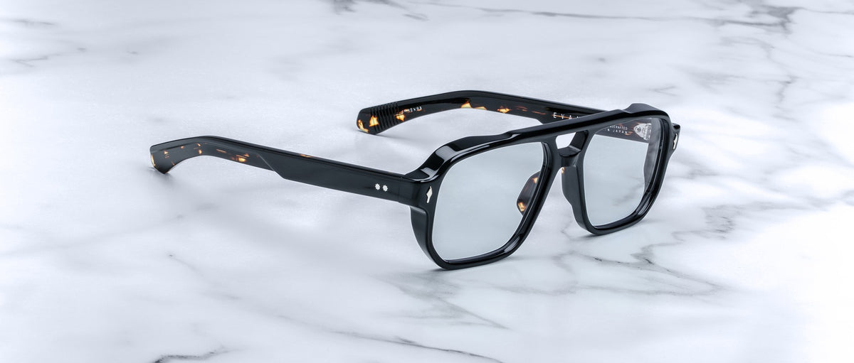 The Jacques Marie Mage Evans eyeglasses feature thick, oversized black acetate frames with clear lenses, echoing classic handcrafted designs, and are displayed on a white marble surface.