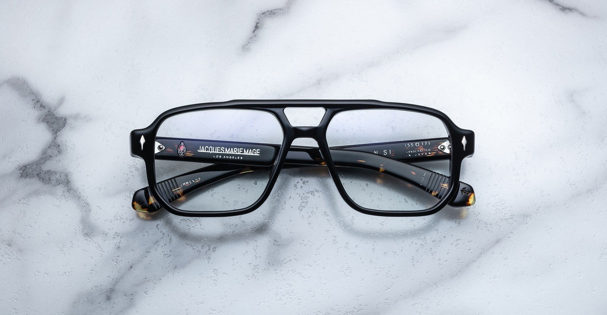 Jacques Marie Mage Evans by Jacques Marie Mage—black square eyeglasses with an oversized acetate frame and clear lenses, arms folded on a white marble surface—a modern handcrafted optical design.
