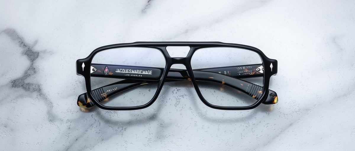Jacques Marie Mage Evans by Jacques Marie Mage—black square eyeglasses with an oversized acetate frame and clear lenses, arms folded on a white marble surface—a modern handcrafted optical design.