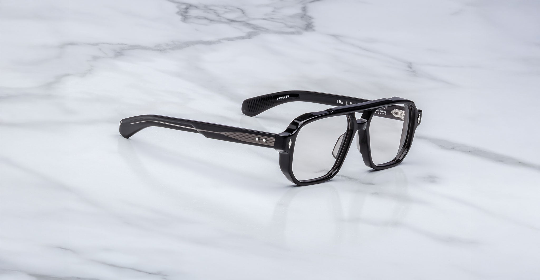 A pair of Jacques Marie Mage Evans black rectangular handcrafted optical glasses with clear lenses rests on a white marble surface.
