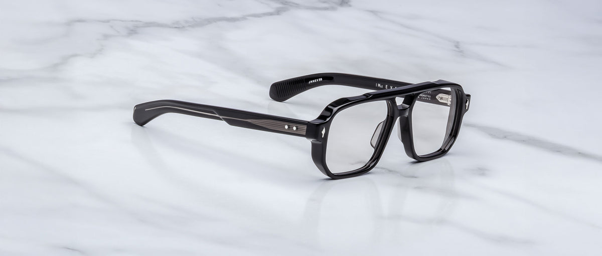 A pair of Jacques Marie Mage Evans black rectangular handcrafted optical glasses with clear lenses rests on a white marble surface.