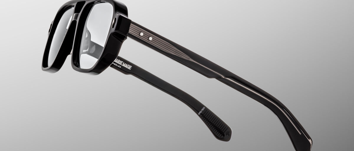 The Jacques Marie Mage Evans are black, thick-rimmed handcrafted optical glasses with clear lenses and metal-accented temples by Jacques Marie Mage, shown against a gray background.