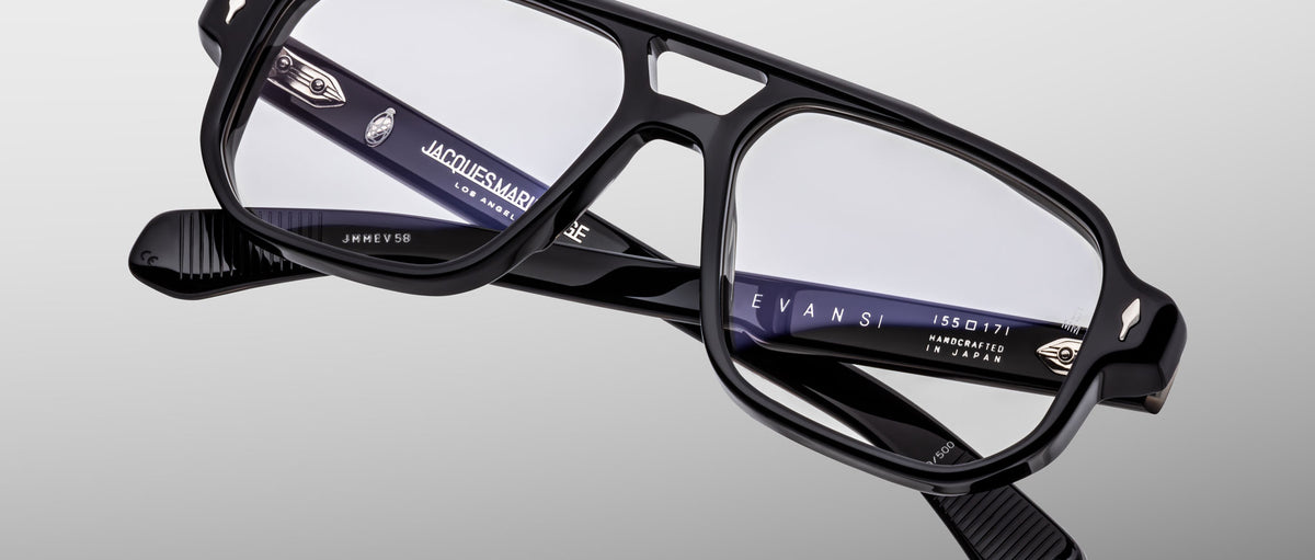 Jacques Marie Mage Evans eyeglass frames in black acetate with an oversized rectangular shape and clear lenses are displayed against a light gray background, featuring visible inner branding on the temples.