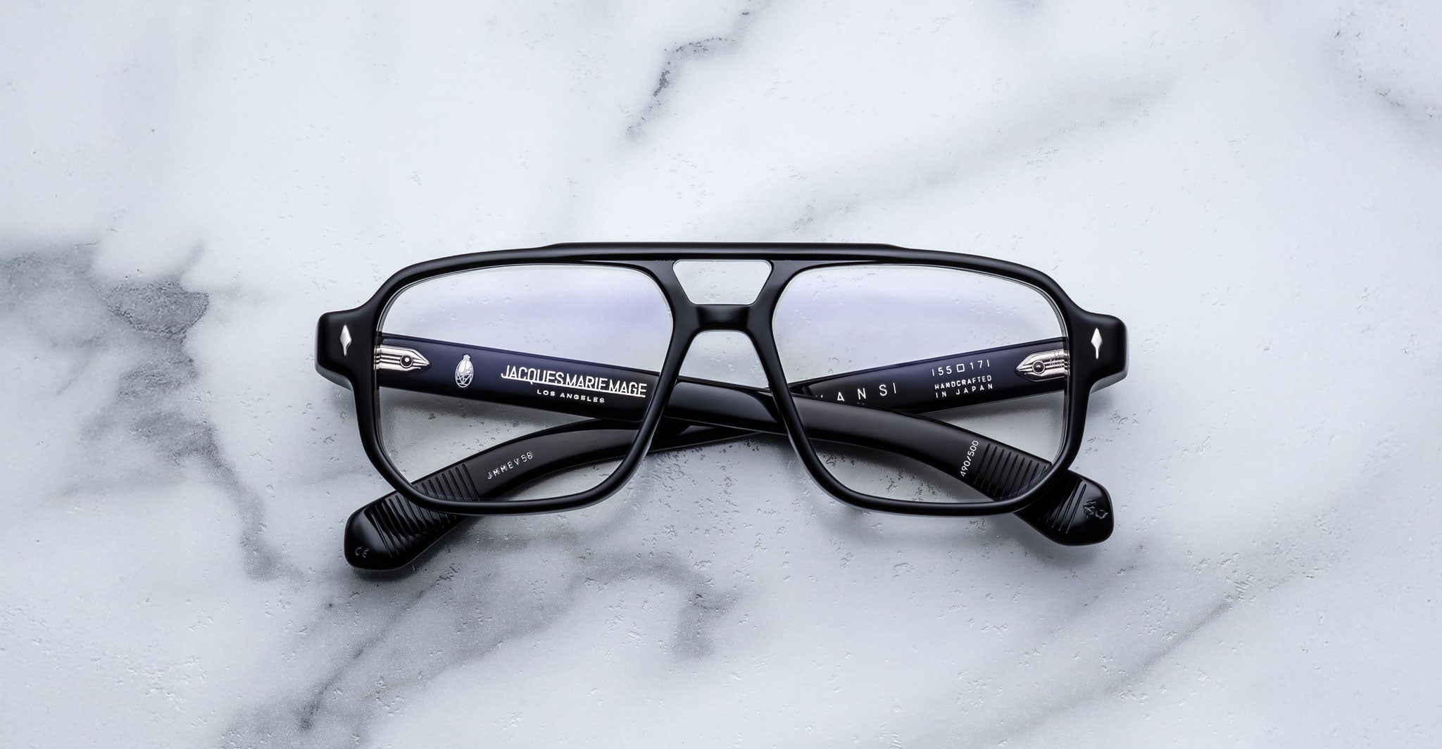 The Jacques Marie Mage Evans by Jacques Marie Mage are black, rectangular, handcrafted optical glasses with clear lenses and an oversized acetate frame, displayed on a white marble surface.