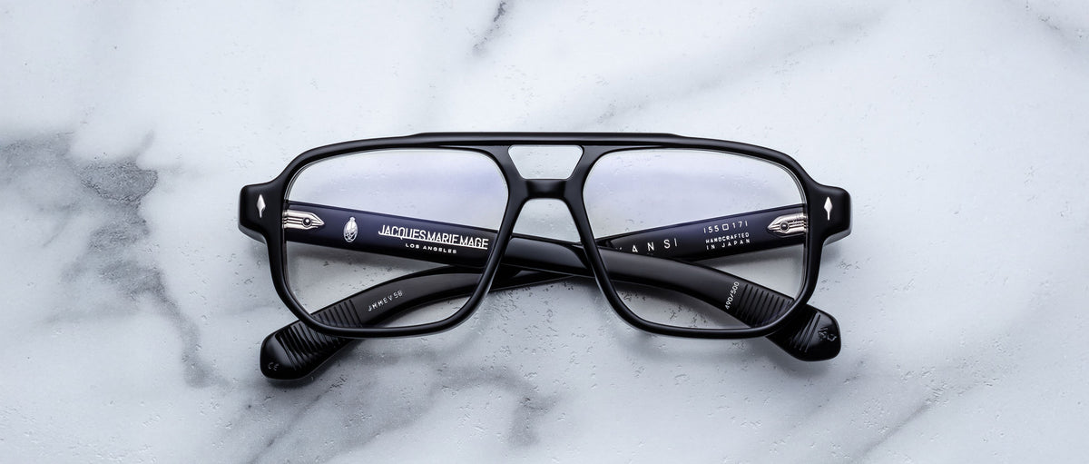 The Jacques Marie Mage Evans by Jacques Marie Mage are black, rectangular, handcrafted optical glasses with clear lenses and an oversized acetate frame, displayed on a white marble surface.