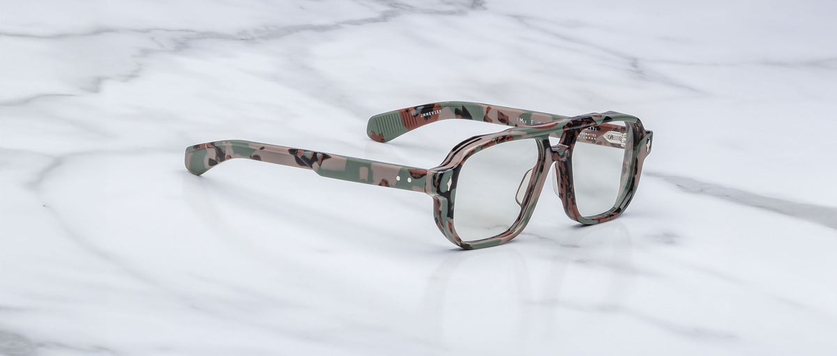 A pair of oversized Jacques Marie Mage Evans eyeglasses with a camouflage-patterned acetate frame and clear lenses rests on a white marble surface.