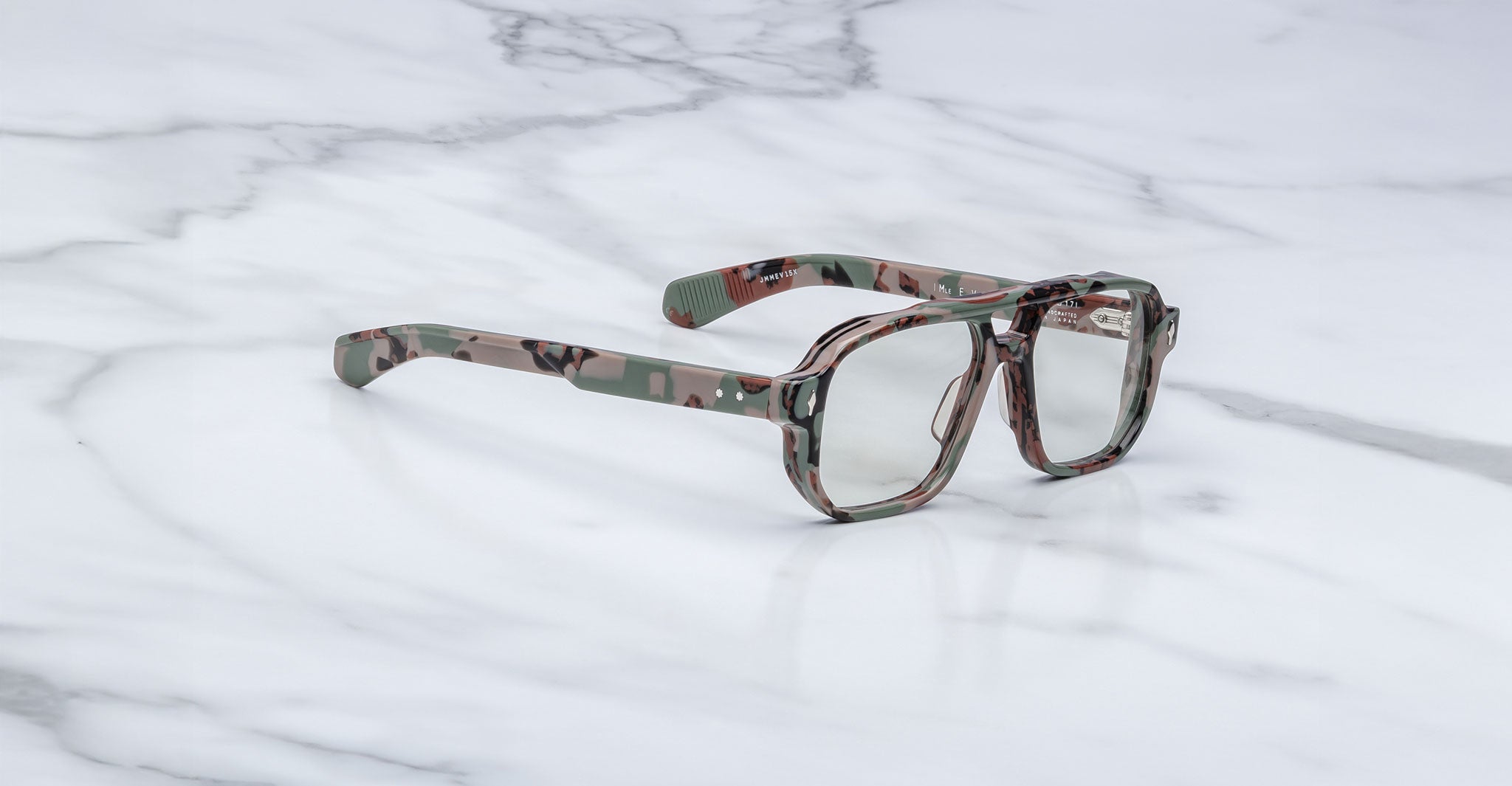 A pair of oversized Jacques Marie Mage Evans eyeglasses with a camouflage-patterned acetate frame and clear lenses rests on a white marble surface.