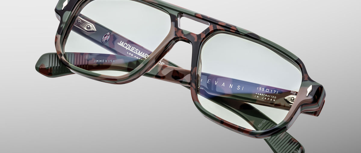 Close-up of Jacques Marie Mage Evans optical glasses by Jacques Marie Mage, featuring a rectangular camouflage design, clear lenses, metallic arm details, and an oversized acetate frame set against a light gray background.