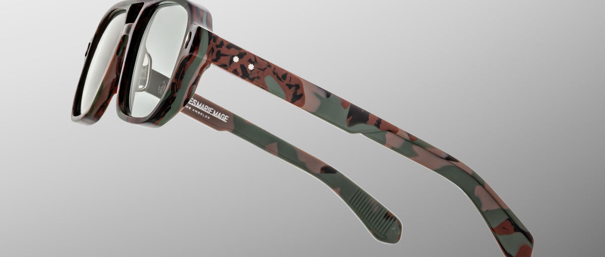 Close-up of Jacques Marie Mage Evans glasses, showcasing handcrafted oversized acetate frames in a camouflage pattern with clear lenses, set against a plain gray background.