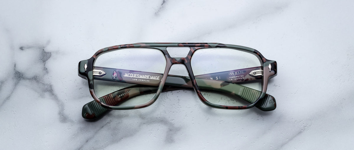 The Jacques Marie Mage Evans optical glasses by Jacques Marie Mage showcase an oversized green camouflage acetate frame with clear lenses, elegantly displayed on a white marble surface.