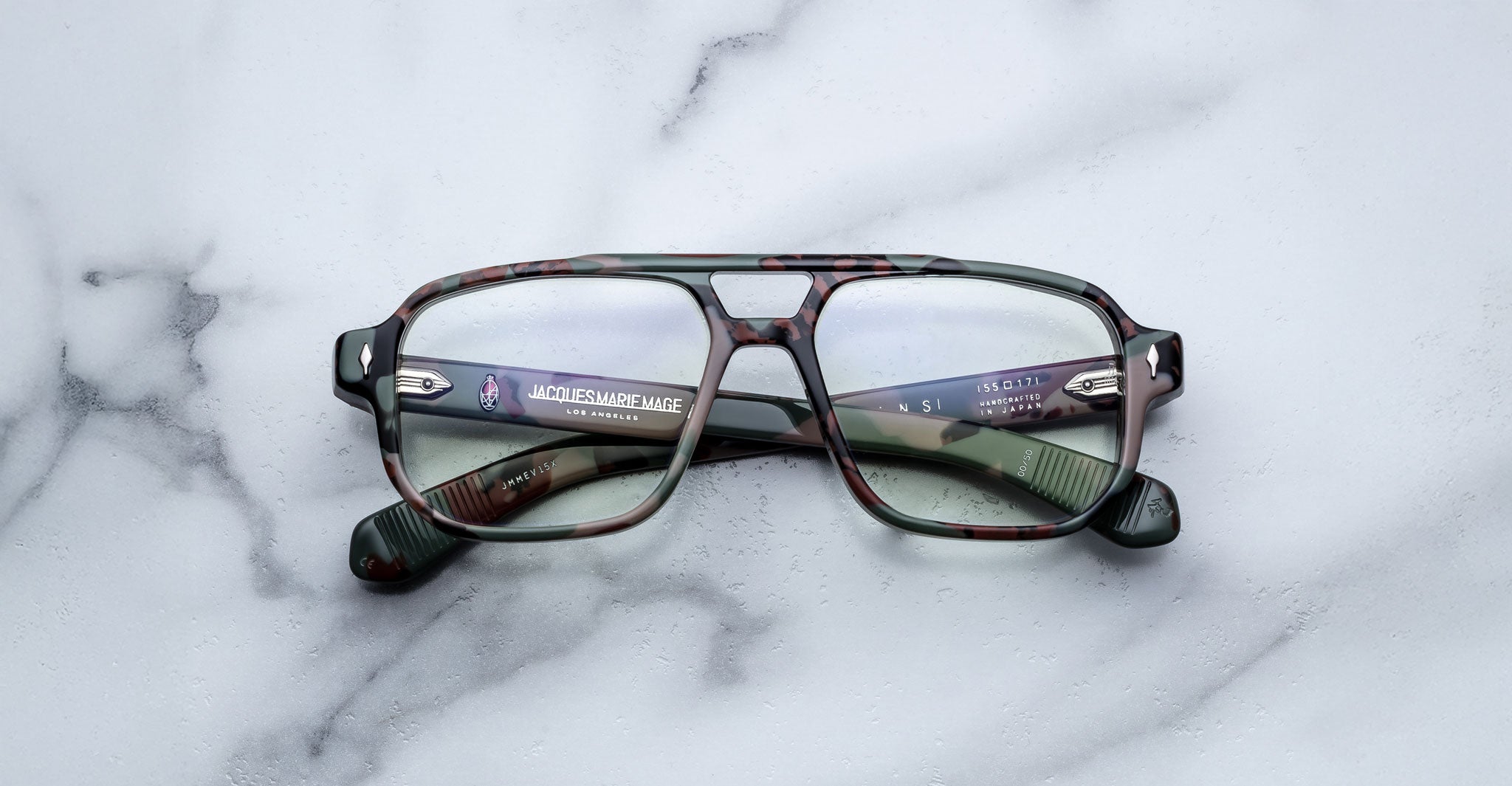 The Jacques Marie Mage Evans optical glasses by Jacques Marie Mage showcase an oversized green camouflage acetate frame with clear lenses, elegantly displayed on a white marble surface.
