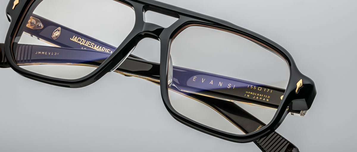 Close-up of Jacques Marie Mage Evans black-rimmed eyeglasses with oversized acetate frames, featuring visible branding on the arms, set against a light grey background.