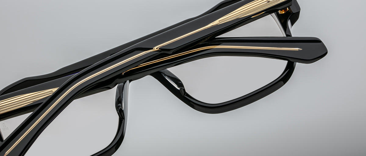 Close-up of Jacques Marie Mage Evans optical glasses, handcrafted with black and gold accents and an oversized acetate frame, shown folded against a plain gray background.