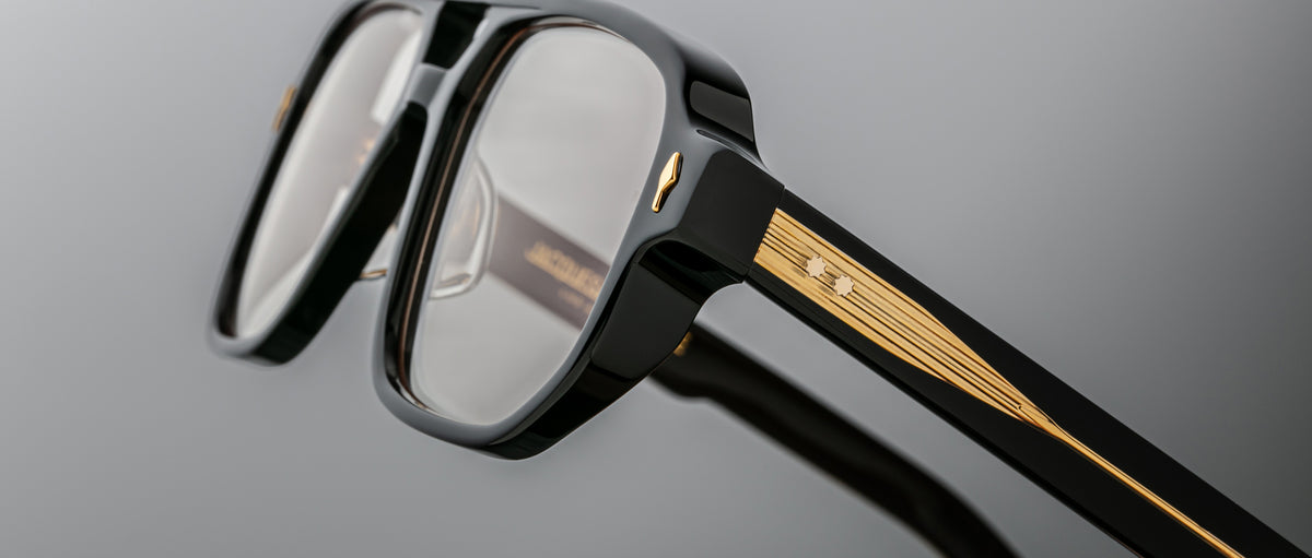 Close-up of Jacques Marie Mage Evans black oversized acetate eyeglasses featuring gold detailing and small star accents on the temples, set against a gray background.