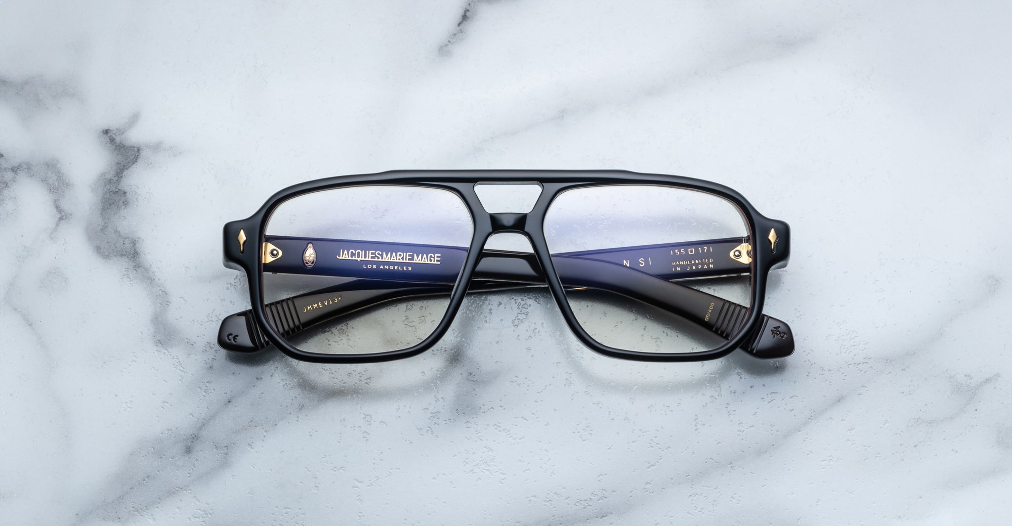 The Jacques Marie Mage Evans optical glasses, featuring black oversized acetate frames and blue inner arms, are displayed on a white marble surface.