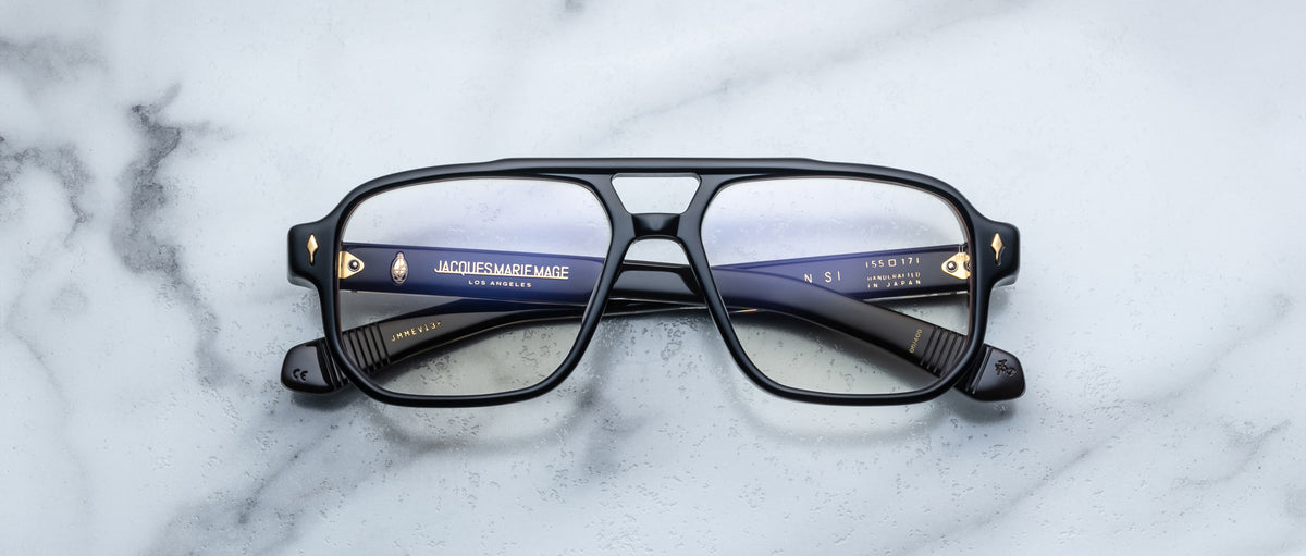 The Jacques Marie Mage Evans optical glasses, featuring black oversized acetate frames and blue inner arms, are displayed on a white marble surface.