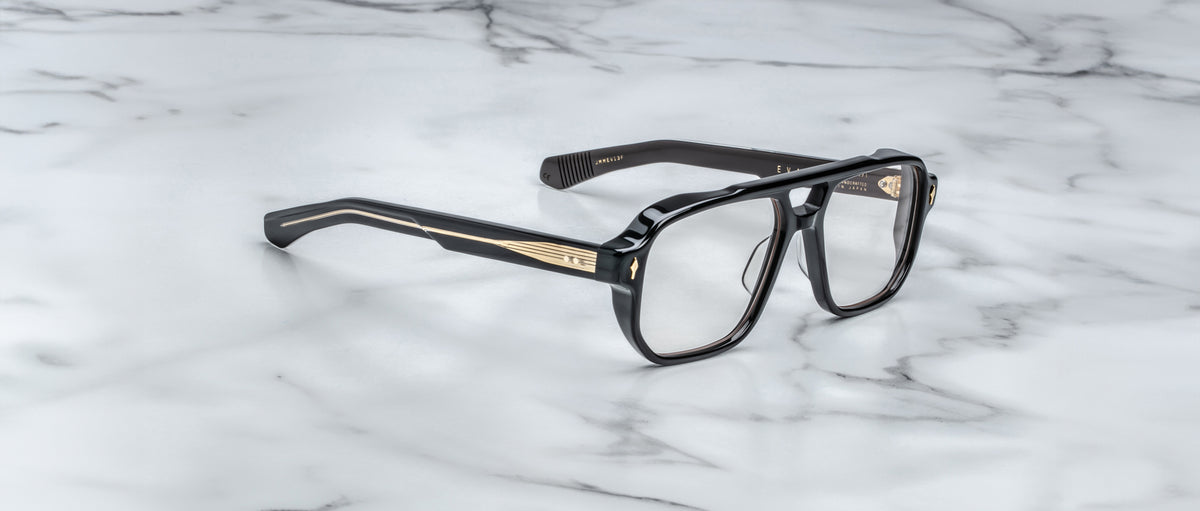 The Jacques Marie Mage Evans handcrafted optical glasses feature an oversized black acetate frame with gold temple detailing, displayed on a white marble surface.