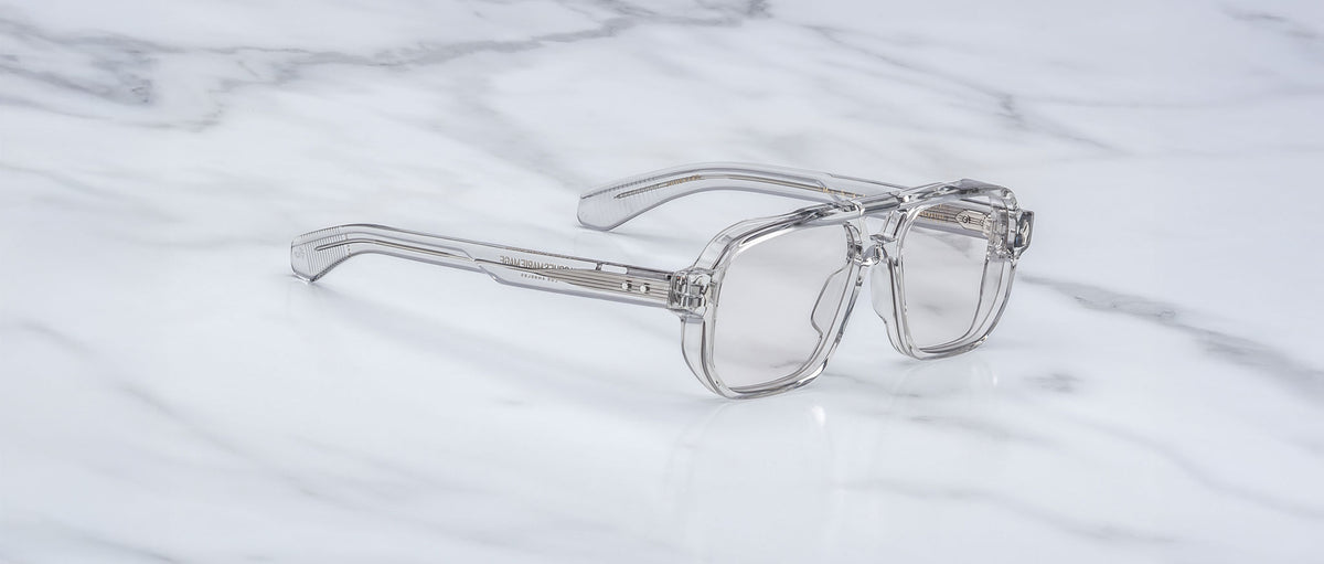 Jacques Marie Mage Evans clear-framed handcrafted optical glasses with transparent lenses are displayed on a white marble surface.