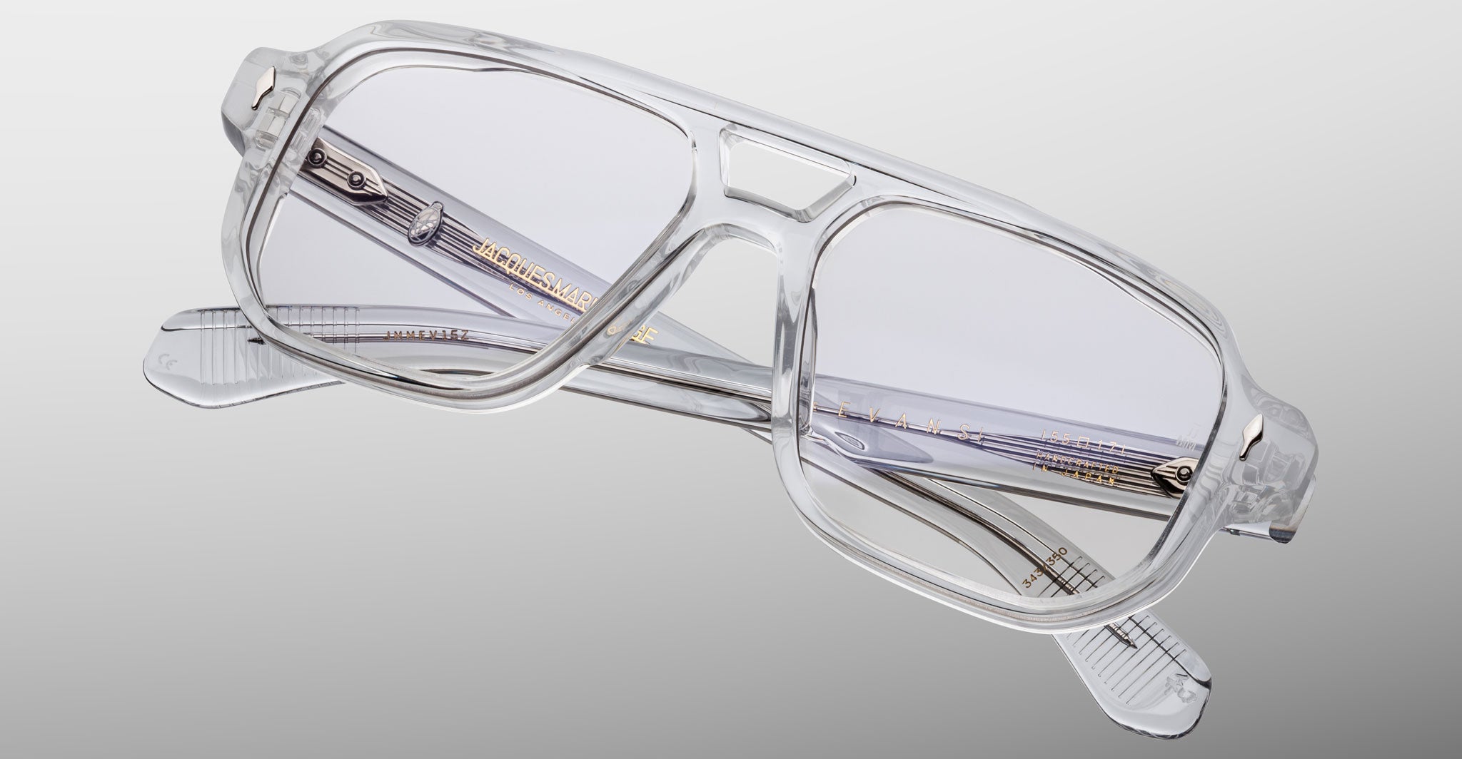 The Jacques Marie Mage Evans by Jacques Marie Mage features transparent rectangular eyeglasses with an oversized acetate frame and gold detailing on the arms, displayed against a light gray background.