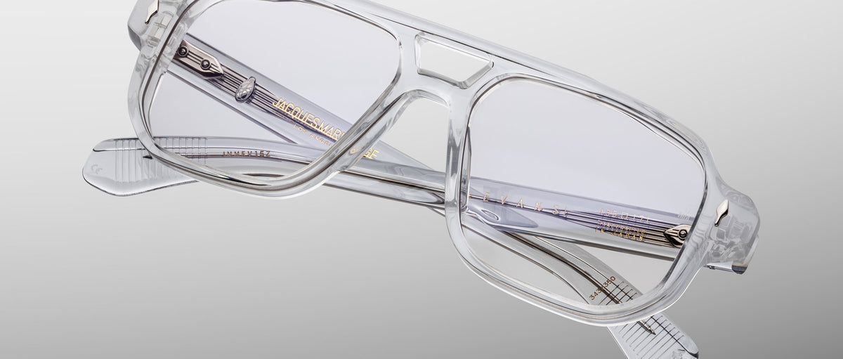 The Jacques Marie Mage Evans by Jacques Marie Mage features transparent rectangular eyeglasses with an oversized acetate frame and gold detailing on the arms, displayed against a light gray background.