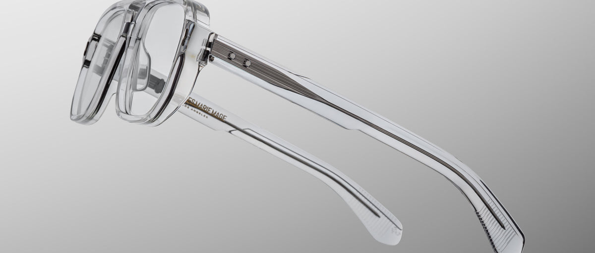 The Jacques Marie Mage Evans optical glasses feature oversized clear acetate frames with translucent arms and visible metal hinges, shown against a neutral gradient background.