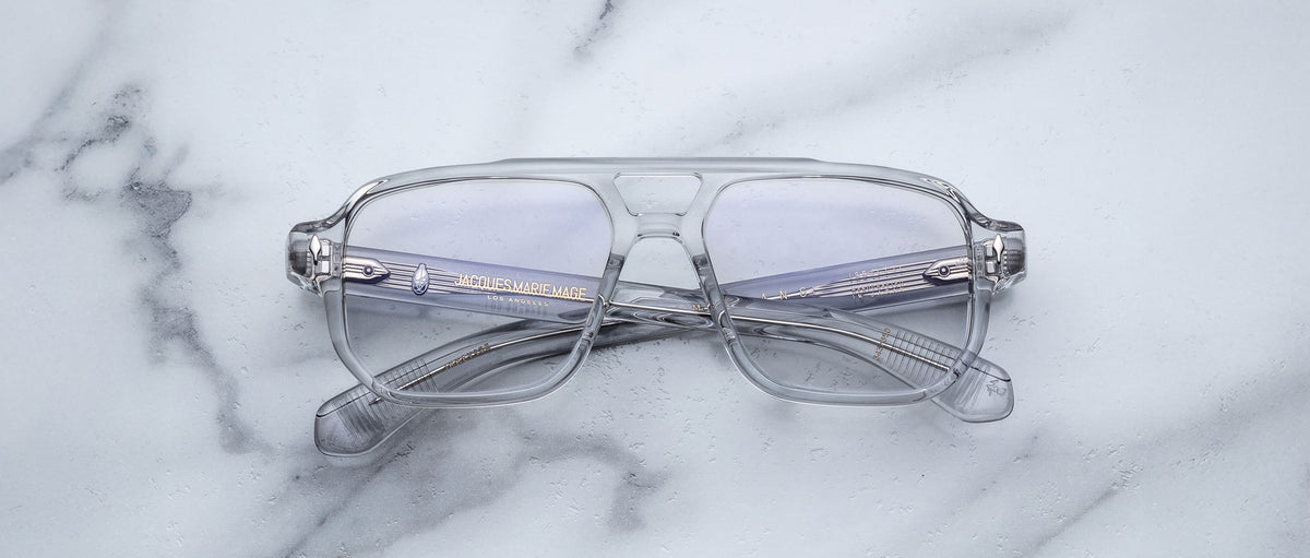 The Jacques Marie Mage Evans by Jacques Marie Mage are oversized acetate eyeglasses with clear frames and slightly tinted lenses, shown folded on a white marble surface.