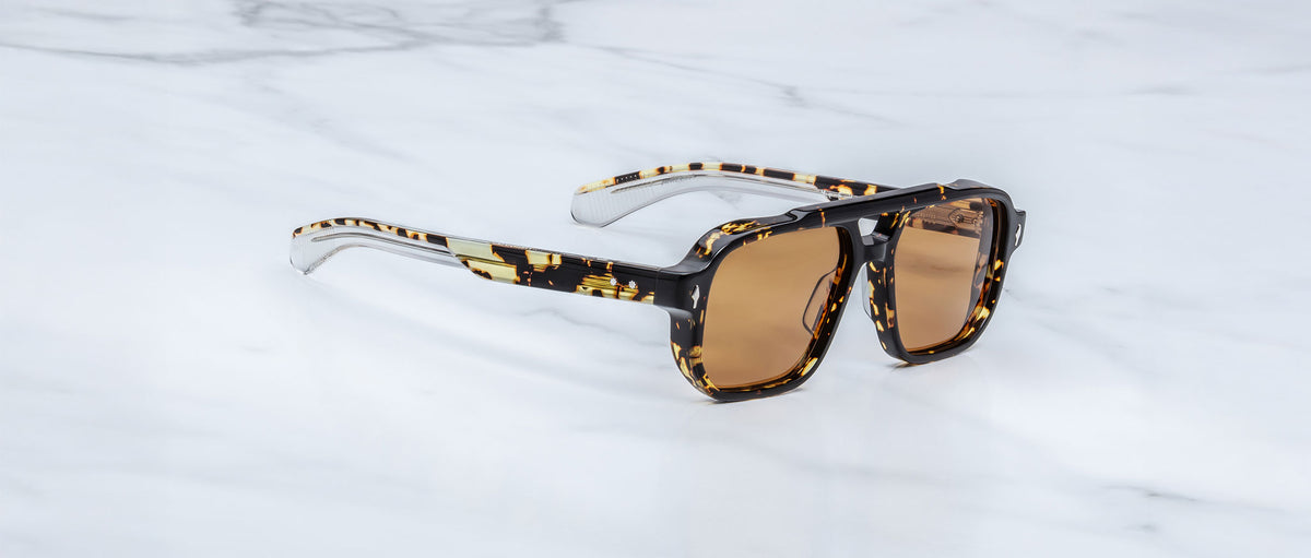 The Jacques Marie Mage Evans sunglasses feature an oversized tortoiseshell acetate frame, brown lenses, and clear arm tips. Displayed on white marble, they embody the refined style of Jacques Marie Mage.
