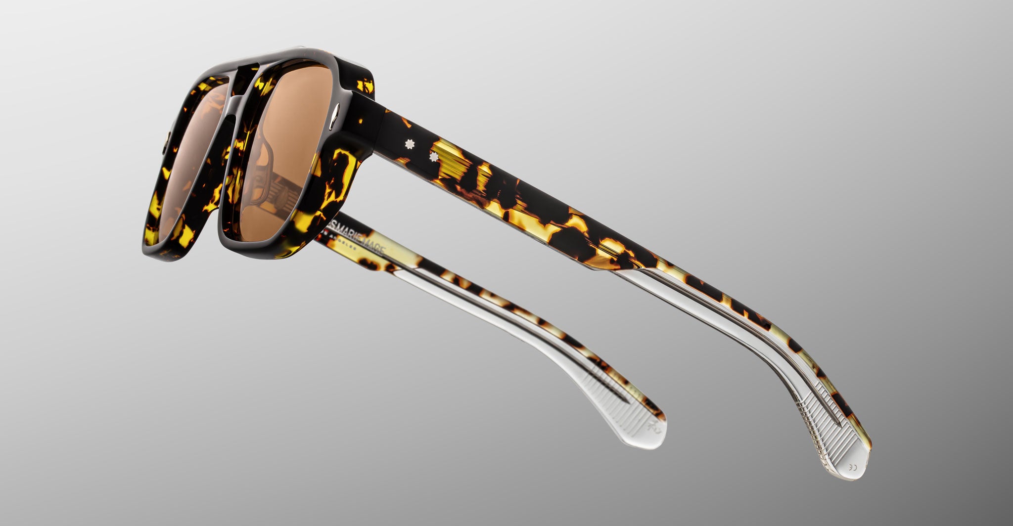 The Jacques Marie Mage Evans sunglasses feature an oversized tortoiseshell acetate frame, brown lenses, and transparent lower arms, displayed against a gradient grey background.