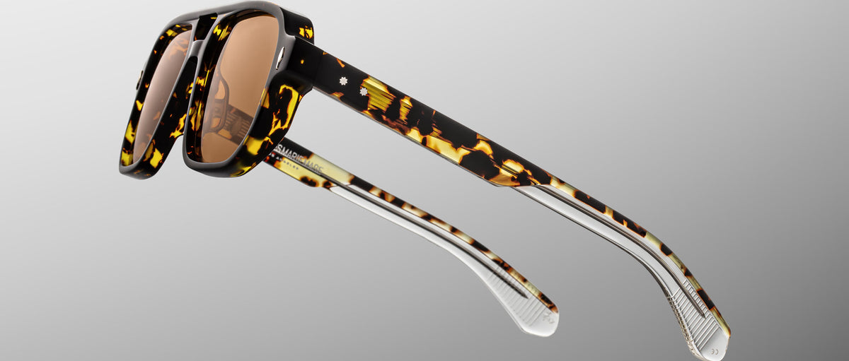 The Jacques Marie Mage Evans sunglasses feature an oversized tortoiseshell acetate frame, brown lenses, and transparent lower arms, displayed against a gradient grey background.