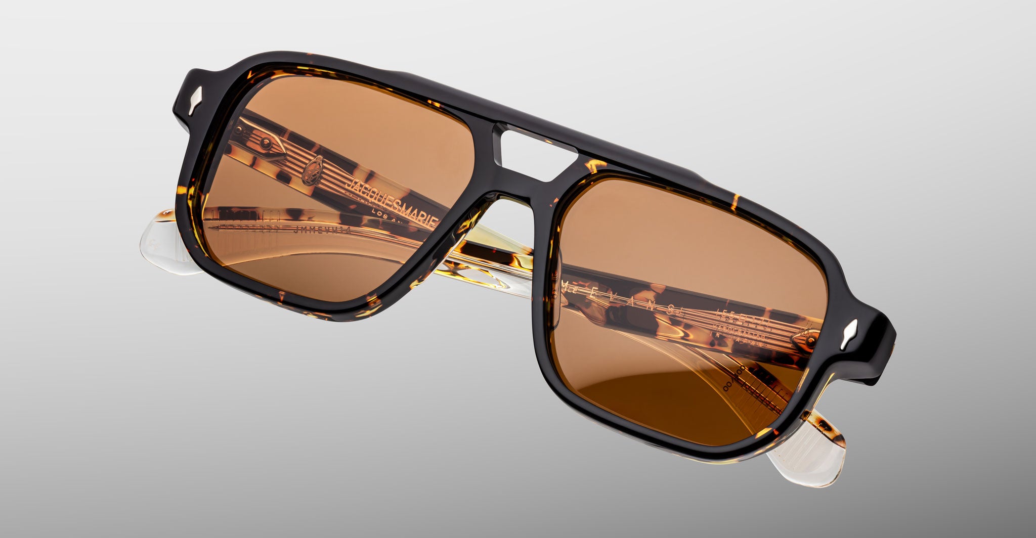 The Jacques Marie Mage Evans sunglasses feature an oversized brown-tinted acetate frame with black and tortoiseshell pattern, gold detailing, and clear temple tips, displayed against a plain gradient background.