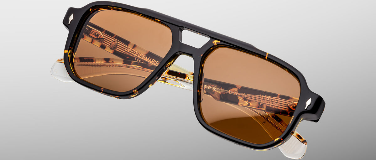 The Jacques Marie Mage Evans sunglasses feature an oversized brown-tinted acetate frame with black and tortoiseshell pattern, gold detailing, and clear temple tips, displayed against a plain gradient background.