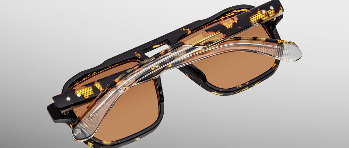 A pair of Jacques Marie Mage Evans sunglasses, featuring an oversized tortoiseshell acetate frame, brown lenses, and clear foldable arms, is folded and viewed from above against a light gray background.