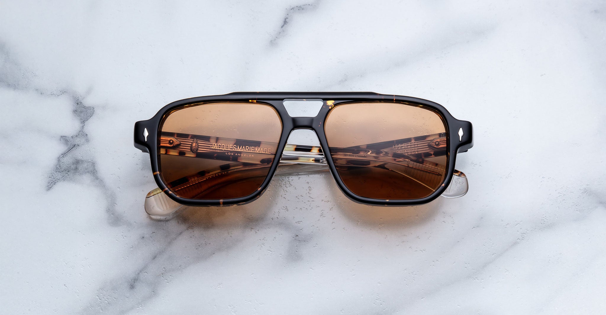A pair of Jacques Marie Mage Evans aviator sunglasses with oversized acetate frames rests on a white marble surface, musical notes pattern reflected in the brown-tinted lenses.