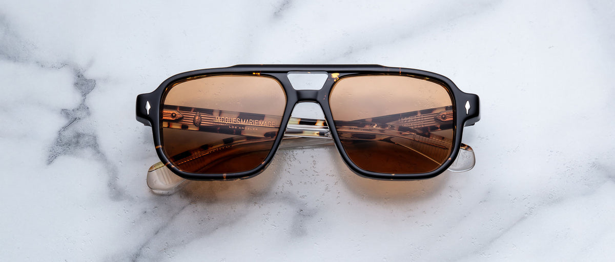 A pair of Jacques Marie Mage Evans aviator sunglasses with oversized acetate frames rests on a white marble surface, musical notes pattern reflected in the brown-tinted lenses.