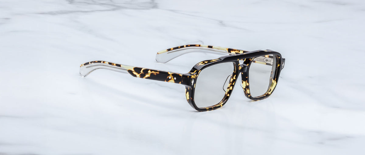 The Jacques Marie Mage Evans are handcrafted optical glasses with oversized tortoiseshell acetate frames and clear lenses, inspired by Jacques Marie Mage, shown resting on a white marble surface.